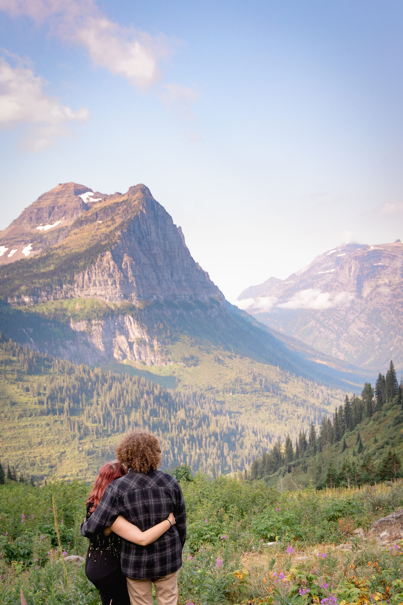 Couple embracing from behind, looking out over a forested valley toward mountain peaks