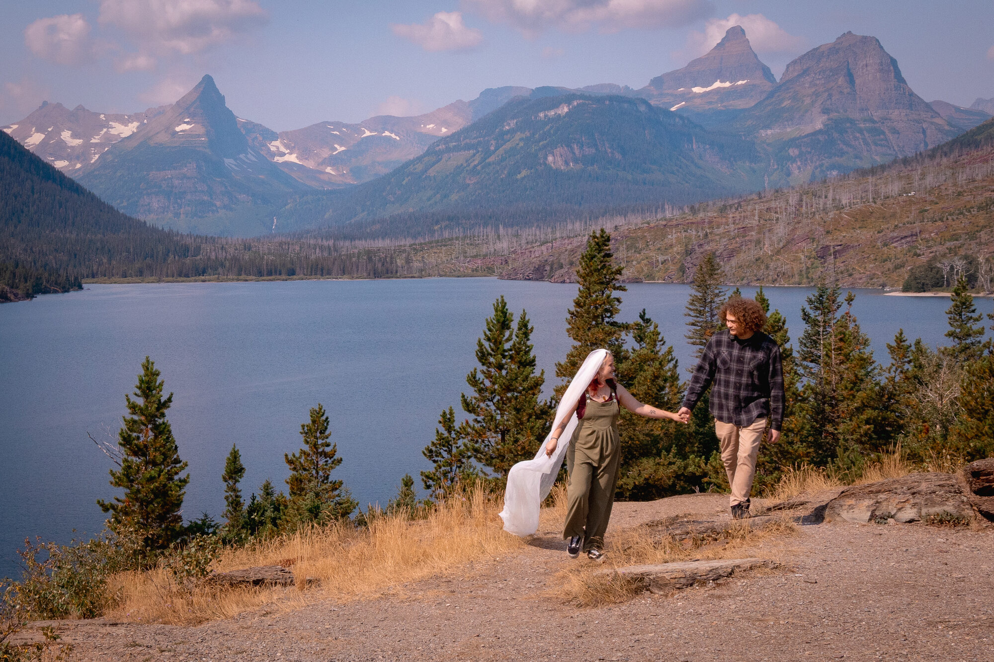 Bride in a flowing veil walking hand in hand with her partner beside an alpine lake